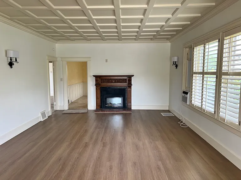 Living Room - featuring original coffered ceiling, new LVT flooring, plantation shutters, Upgraded LED electric fireplace, original carved wood mantel and window A/C unit.
