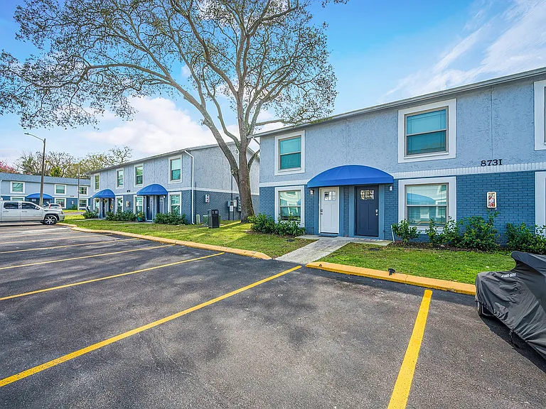 Charming apartment building with blue awnings and lush greenery, featuring ample parking on a sunny day.