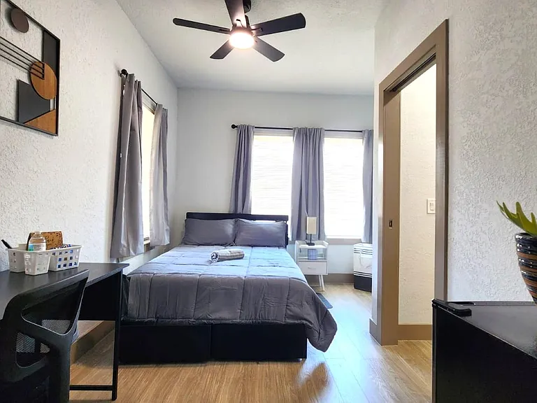 Furnished bedroom with queen bed, desk workspace, and dual windows with curtains, plus a ceiling fan and nightstand.