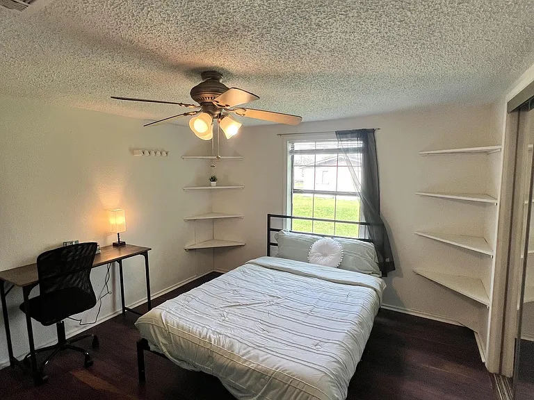 Furnished bedroom with a ceiling fan, work desk, and built-in wall shelving, plus a window with natural light.