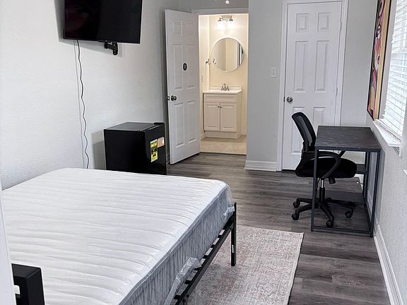 Private furnished bedroom with a bed, wall-mounted TV, and desk area, plus convenient access to a vanity sink just outside.