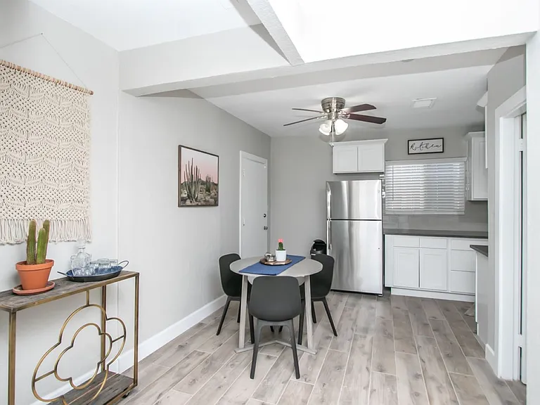 Detached casita dining and kitchen area at Tempe rental home. Features a round dining table with modern black chairs, a stainless steel refrigerator, white shaker cabinets, wood ceramic flooring, desert wall art. Open layout for a top Tempe, AZ stay.