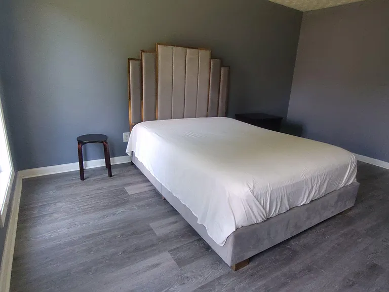 Spacious bedroom with a large bed, tall headboard, and durable wood-look flooring, plus a window for natural light.