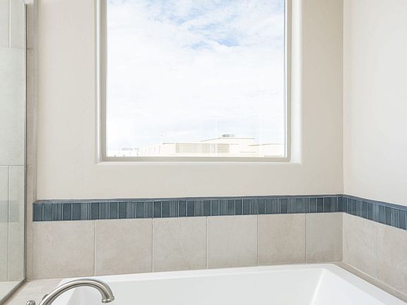 A modern white bathtub with silver fixtures is set against beige tiled walls. A large window above t