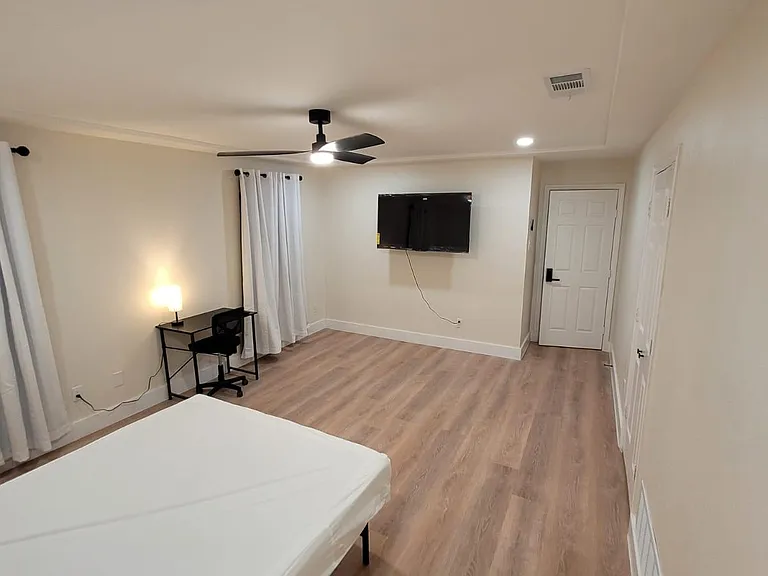 Spacious bedroom with wood-look flooring, ceiling fan, wall-mounted TV, and a dedicated desk area with window curtains.