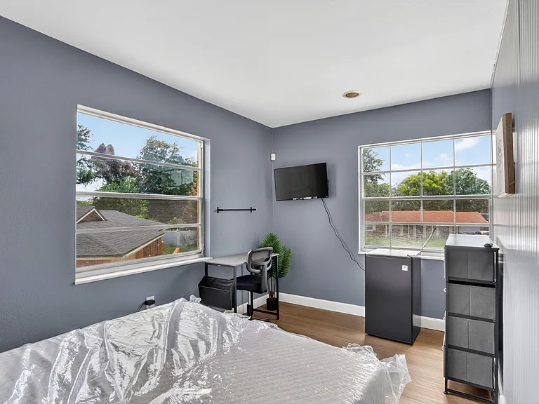 Private bedroom with two large windows, a wall-mounted TV, and space for a small desk setup or storage.
