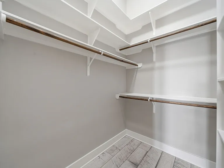 Spacious walk-in closet featuring custom white shelving and warm wood hanging rods, offering exceptional storage and organization. The neutral finishes and light flooring create a bright, open feel.