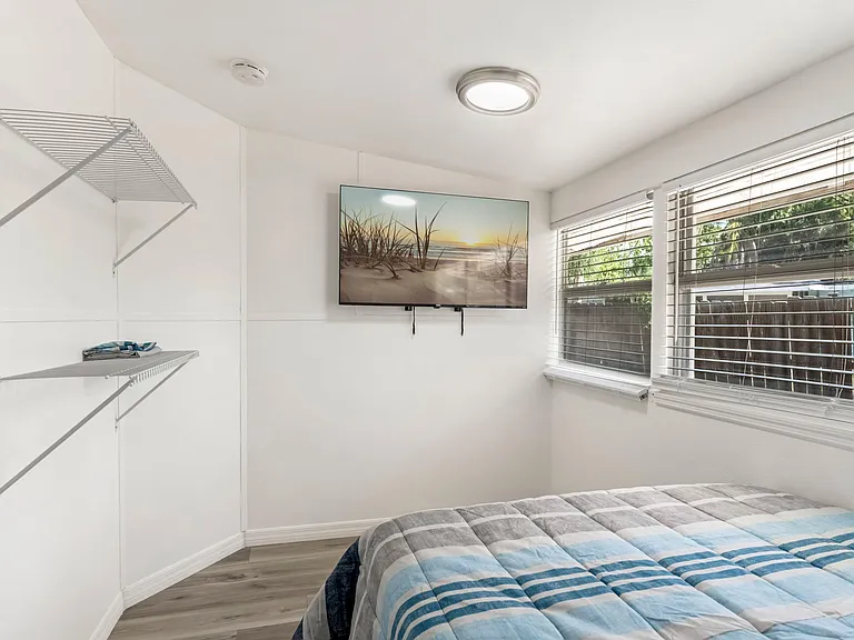 Bedroom with built-in closet shelving, wall-mounted TV, and large window with blinds for natural light and privacy.