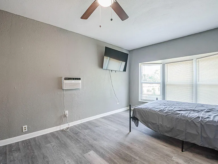 Bedroom with wide windows and blinds, ceiling fan, wall-mounted TV, and durable wood-look flooring with space for furnishings
