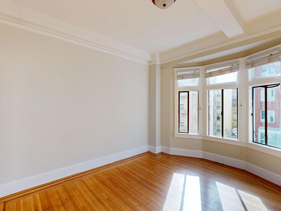 Bright, empty room with hardwood floors and large bay windows, featuring intricate ceiling details.