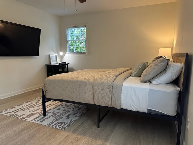 Furnished bedroom with full-size bed, wall-mounted TV, dedicated desk area, and a window with blinds for natural light.