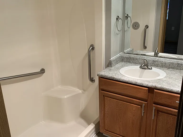 Clean guest bathroom featuring a walk-in shower with safety grab bars, a spacious vanity with ample counter space, and warm wood cabinetry. Bright lighting and a functional layout make it both comfortable and convenient.