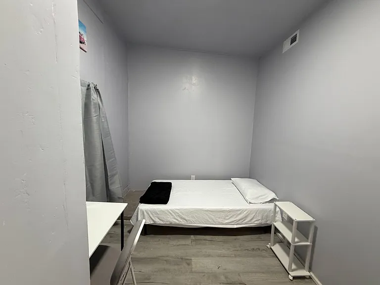 Private bedroom with a single bed, side table, and compact work surface, plus durable flooring and neutral walls.