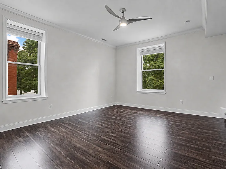 Bright first-floor family room with 10-foot ceilings, two windows with 2-inch wood Venetian blinds, wood-style flooring, crown molding, and a modern ceiling fan.
