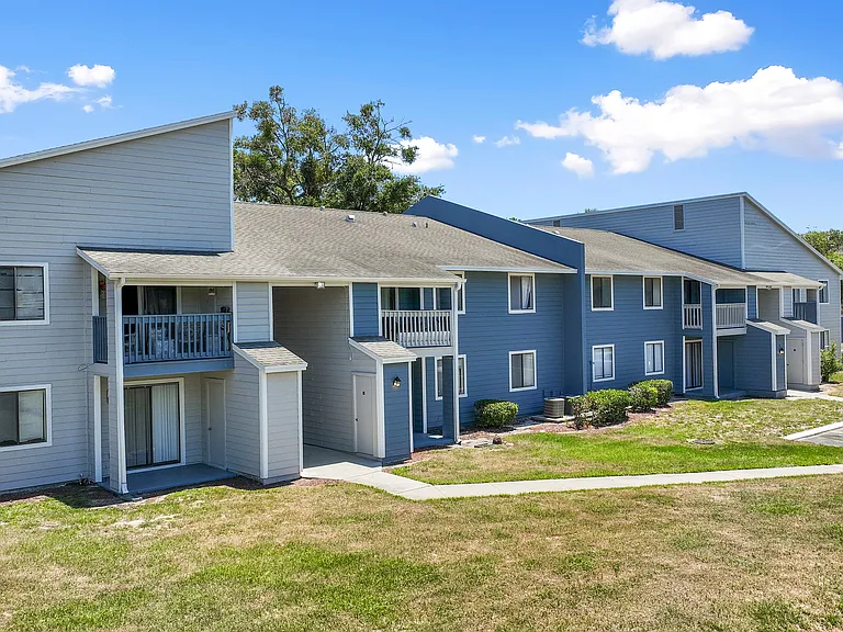 Charming two-story apartment complex with blue siding, nestled amid lush greenery under a bright blue sky.
