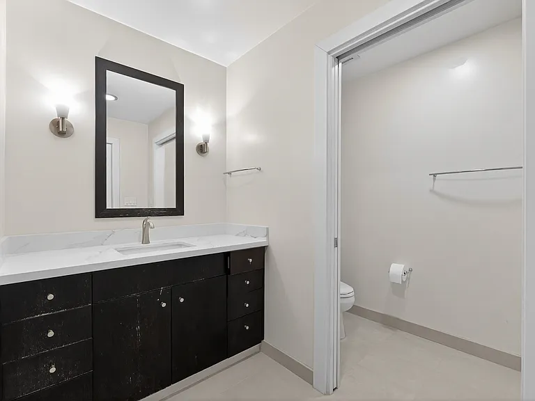 Bathroom vanity with updated countertop, modern faucet, wall sconces, and separate toilet area.