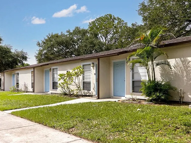 Charming duplex with light blue doors surrounded by lush greenery and mature trees, offering serene living in a peaceful neighborhood.