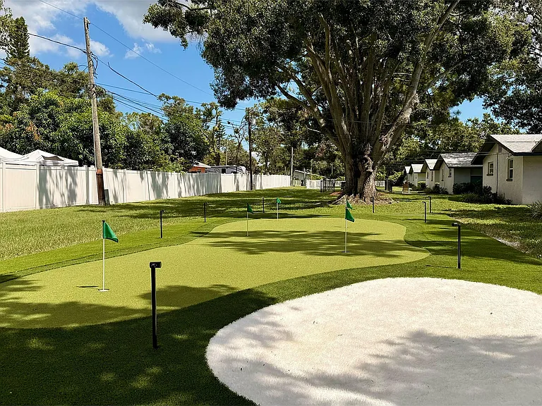 Charming outdoor view with a well-maintained putting green surrounded by lush greenery and quaint cottages under a clear blue sky.