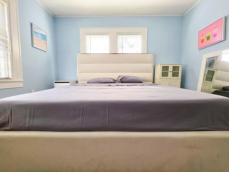 Spacious bedroom with a large bed, dual windows with blinds, and a storage cabinet plus full-length mirror.