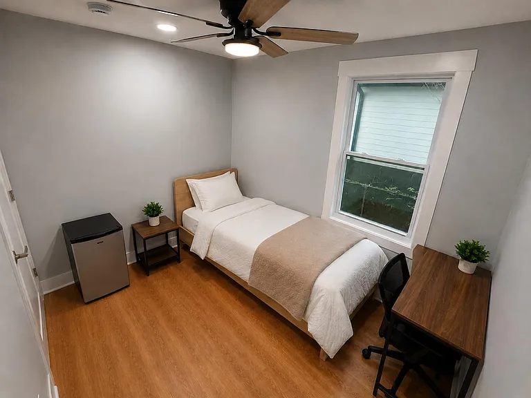 Efficient private bedroom with twin bed, desk workspace, mini fridge, and a window for natural light and ventilation.
