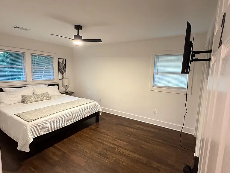 Spacious bedroom with hardwood floors, ceiling fan, and multiple windows; wall-mounted TV adds convenient entertainment.