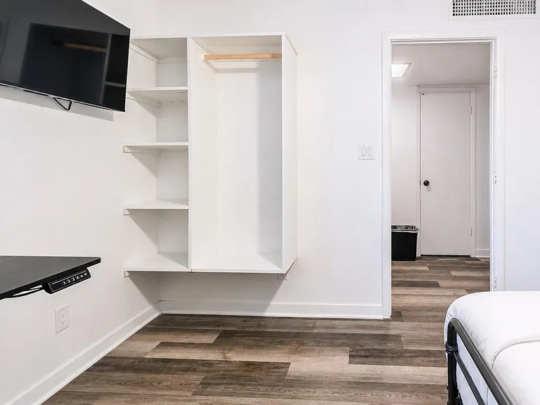 Bedroom features wood-look flooring, wall-mounted TV, and an open closet with shelving and hanging rod for storage.