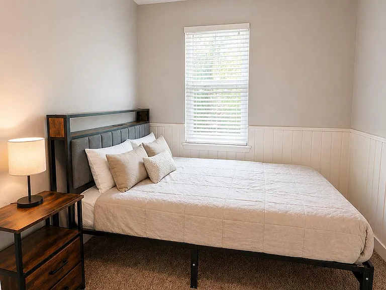 Furnished bedroom featuring a full-size bed, nightstand with lamp, and a window with blinds for natural light.