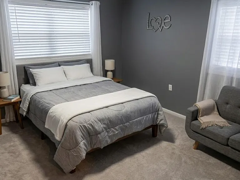 Well-sized bedroom with plush carpet, two windows with blinds and curtains, and space for a sitting area.