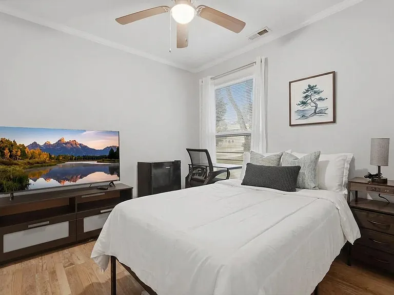 Furnished bedroom with a full bed, nightstand, dresser with TV, desk chair, ceiling fan, and a window with blinds.