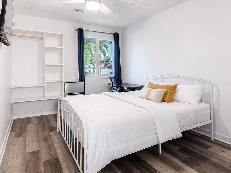 Furnished bedroom with full bed, dedicated desk, open shelving/closet storage, and a window with curtain panels.
