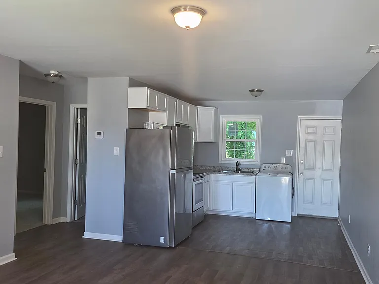 Bright and spacious open-concept kitchen and living area featuring modern gray walls, durable wood-style flooring, and a functional kitchen layout. The space includes white cabinetry, ample counter space, a full-size refrigerator, and a large window that