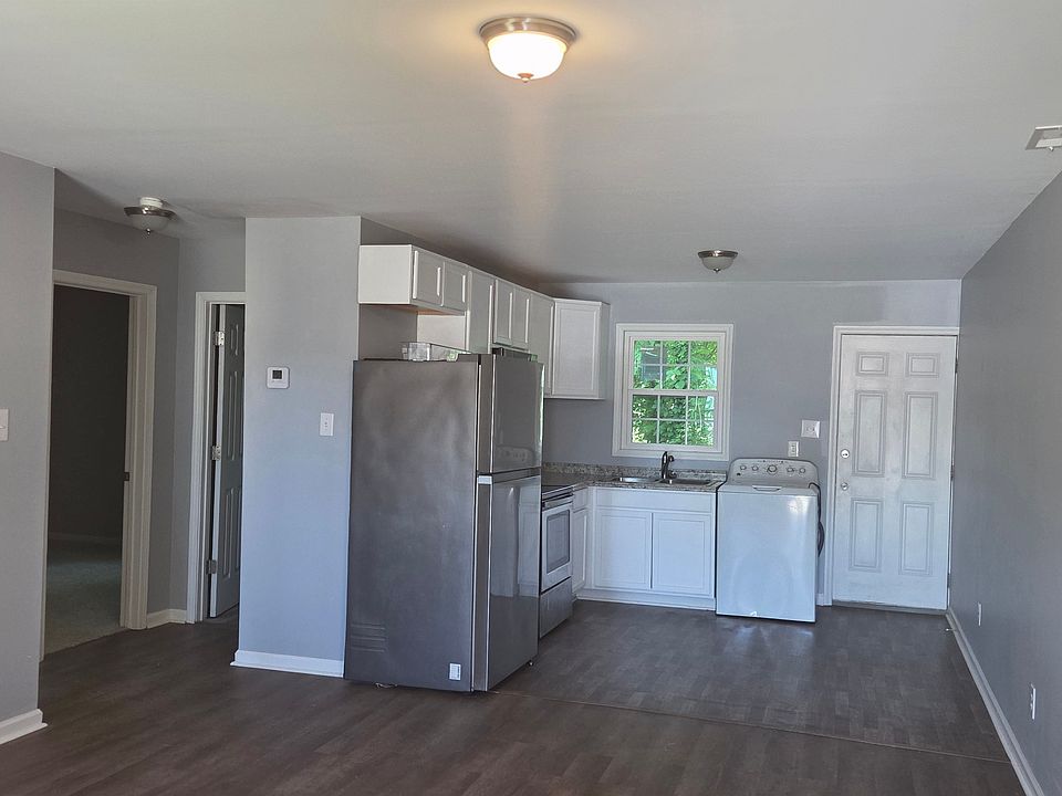 Bright and spacious open-concept kitchen and living area featuring modern gray walls, durable wood-style flooring, and a functional kitchen layout. The space includes white cabinetry, ample counter space, a full-size refrigerator, and a large window that 