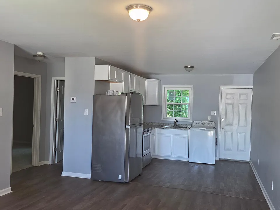 Bright and spacious open-concept kitchen and living area featuring modern gray walls, durable wood-style flooring, and a functional kitchen layout. The space includes white cabinetry, ample counter space, a full-size refrigerator, and a large window that 