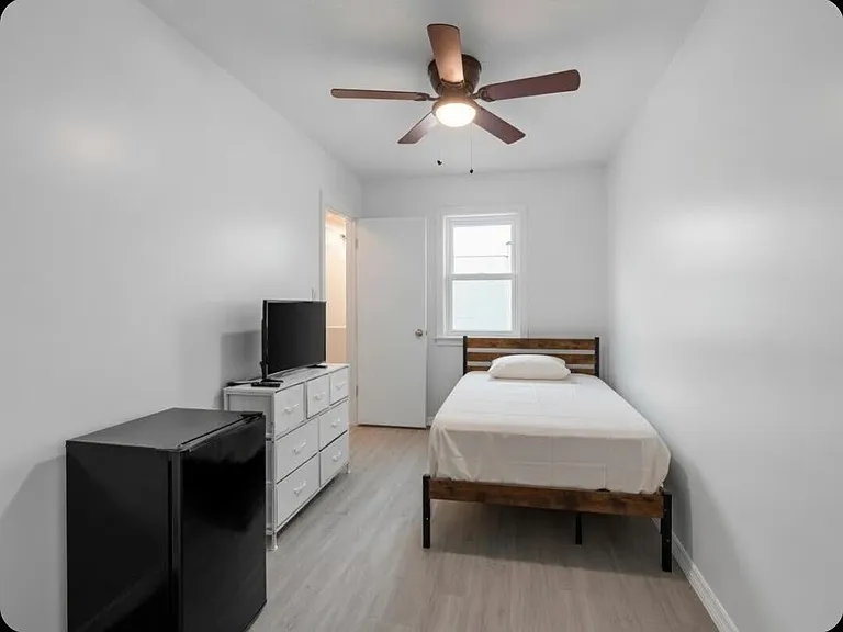 Private bedroom with a ceiling fan, window, and furnished setup including bed frame, dresser, TV stand, and mini fridge.