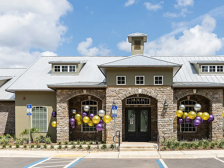 Charming residential building with elegant stone facade and inviting entrance, adorned with colorful balloons, surrounded by lush landscaping under a vibrant blue sky.