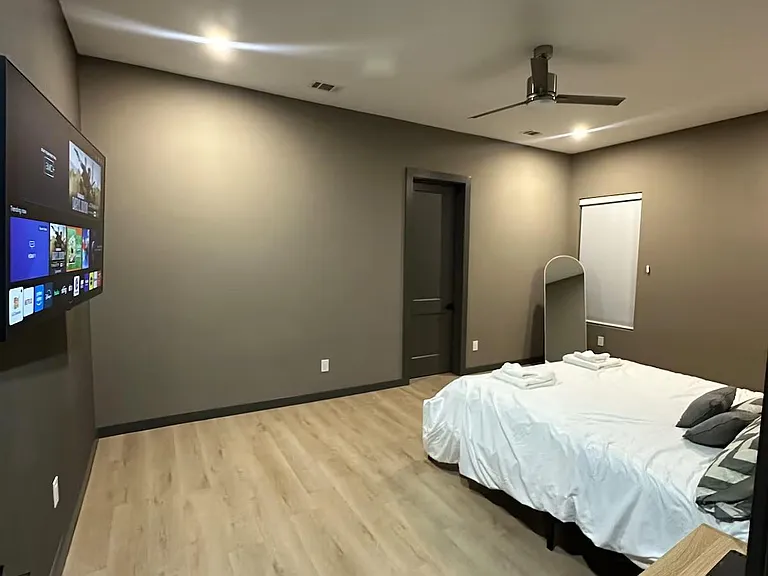 Spacious bedroom with wood-look floors, ceiling fan, mounted TV, and a window with shade for added privacy.