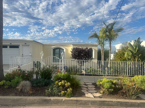 Striking modern facade with arched windows, tropical landscaping, and a white metal fence. A stone path leads through lush succulents and palms to a gated entrance, framed by lantern-style lights_welcoming you with elegance and coastal charm.