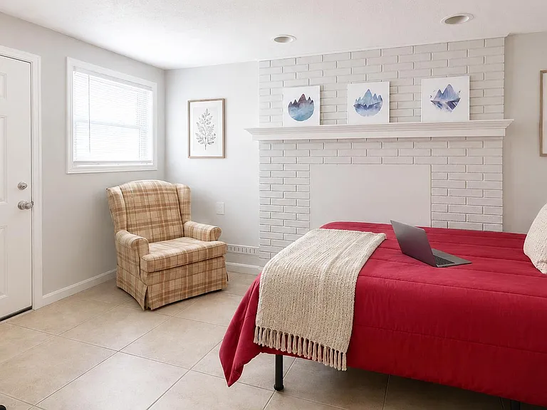 Spacious furnished bedroom with tile flooring, large window, and a decorative brick fireplace feature wall with mantel.