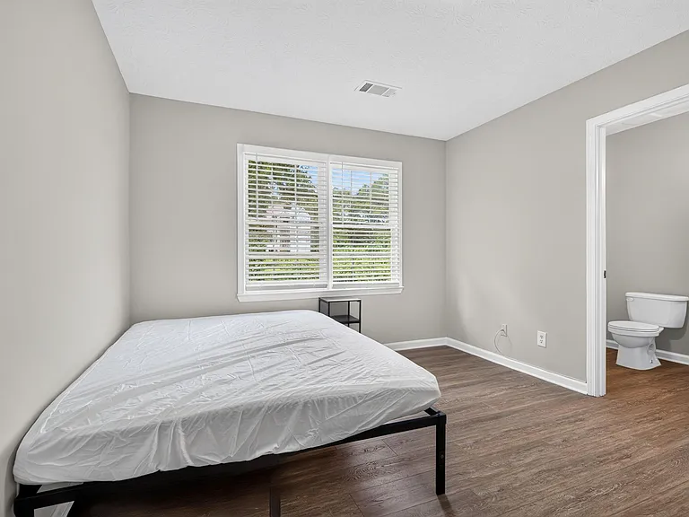 Bedroom with large window and blinds, wood-look flooring, and convenient access to a nearby bathroom.