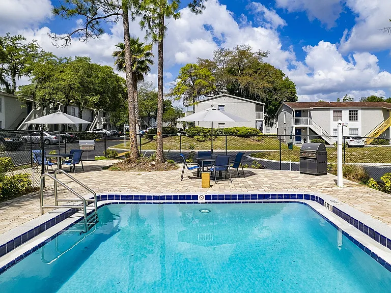 Charming apartment complex featuring a refreshing outdoor pool, relaxing patio furniture, and a barbecue area surrounded by lush greenery and elegant architecture.