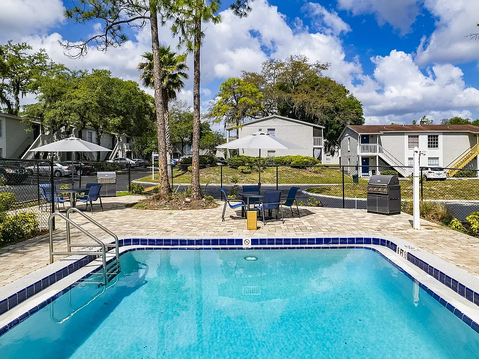 Charming apartment complex featuring a refreshing outdoor pool, relaxing patio furniture, and a barbecue area surrounded by lush greenery and elegant architecture.