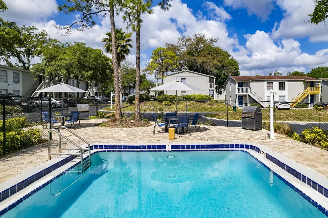 Charming apartment complex featuring a refreshing outdoor pool, relaxing patio furniture, and a barbecue area surrounded by lush greenery and elegant architecture.