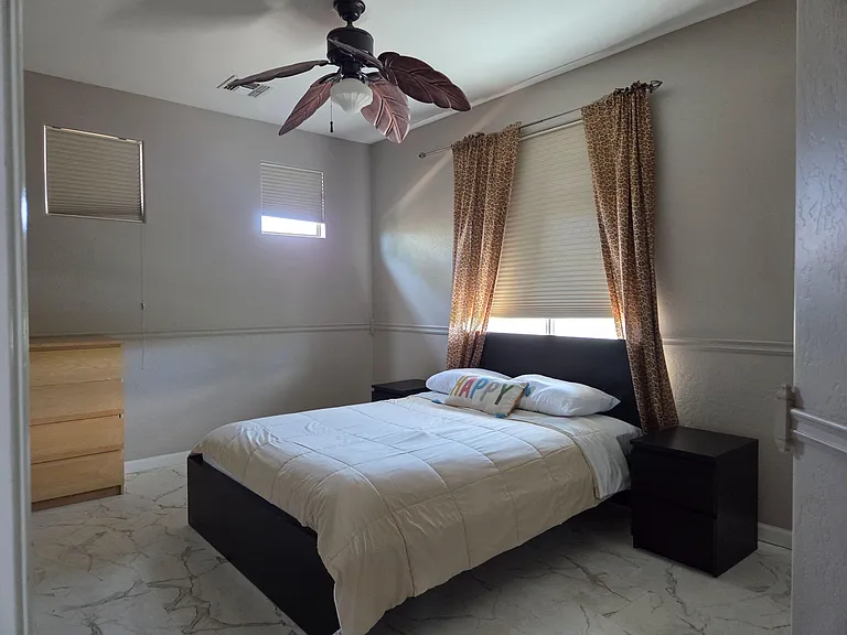 Furnished bedroom featuring a queen bed, nightstands, dresser, and ceiling fan, with multiple windows and window coverings.