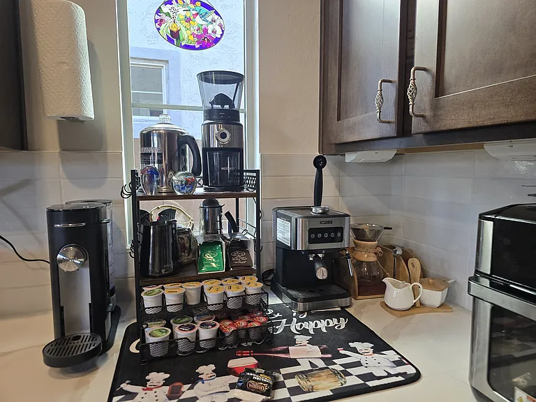 This cozy coffee station features a Ninja brewer, Keurig, and espresso machine with frother, plus a neatly arranged pod selection. Set on white counters with dark wood cabinetry, it's a barista-style setup with natural light and curated charm.