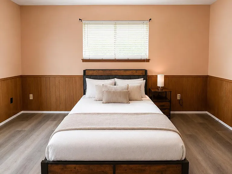 Spacious bedroom featuring a full bed setup, side table with lamp, and a large window with blinds for controlled light.