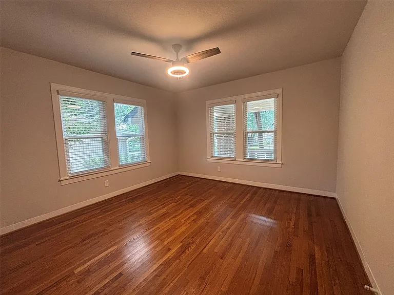 Spacious room featuring rich wood-finish flooring, a contemporary ceiling fan with integrated lighting, and two large windows with blinds