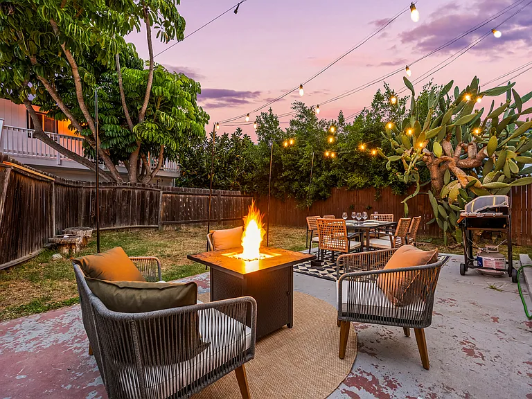 Cozy backyard retreat with cafe-style string lights, outdoor dining, a BBQ grill, and a fire pit lounge. Surrounded by palm trees and warm lighting, this inviting space is perfect for relaxing evenings and group gatherings.
