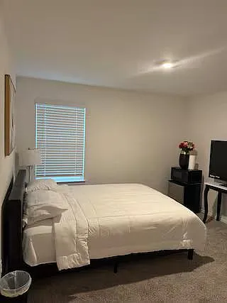Furnished bedroom with a full bed, side tables, and a TV setup, plus window blinds and carpeted flooring.