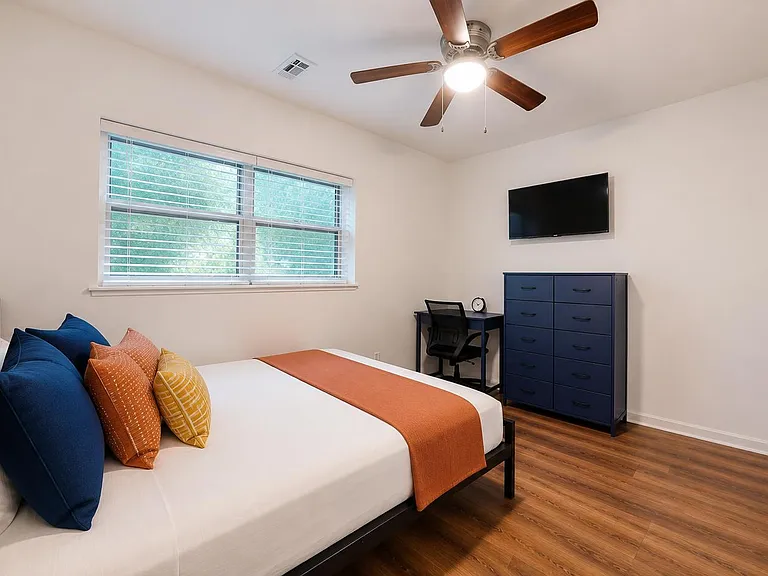 Furnished bedroom with a large window, ceiling fan, wall-mounted TV, dedicated desk, and dresser storage.