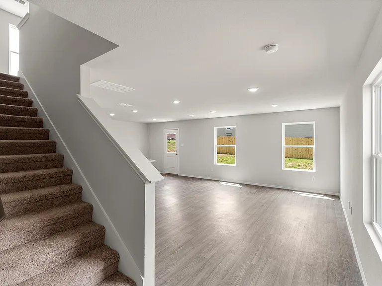 Spacious open-concept living area featuring wood-finish flooring, recessed lighting, multiple windows, and a carpeted staircase with a painted railing